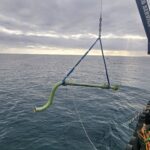 photo of green pipe with flanges and a valve being lifted by a crane over the water's surface