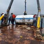 photo of workers on back deck of a boat while large, pill-shaped buoy is lifted onto the deck by stern A-frame